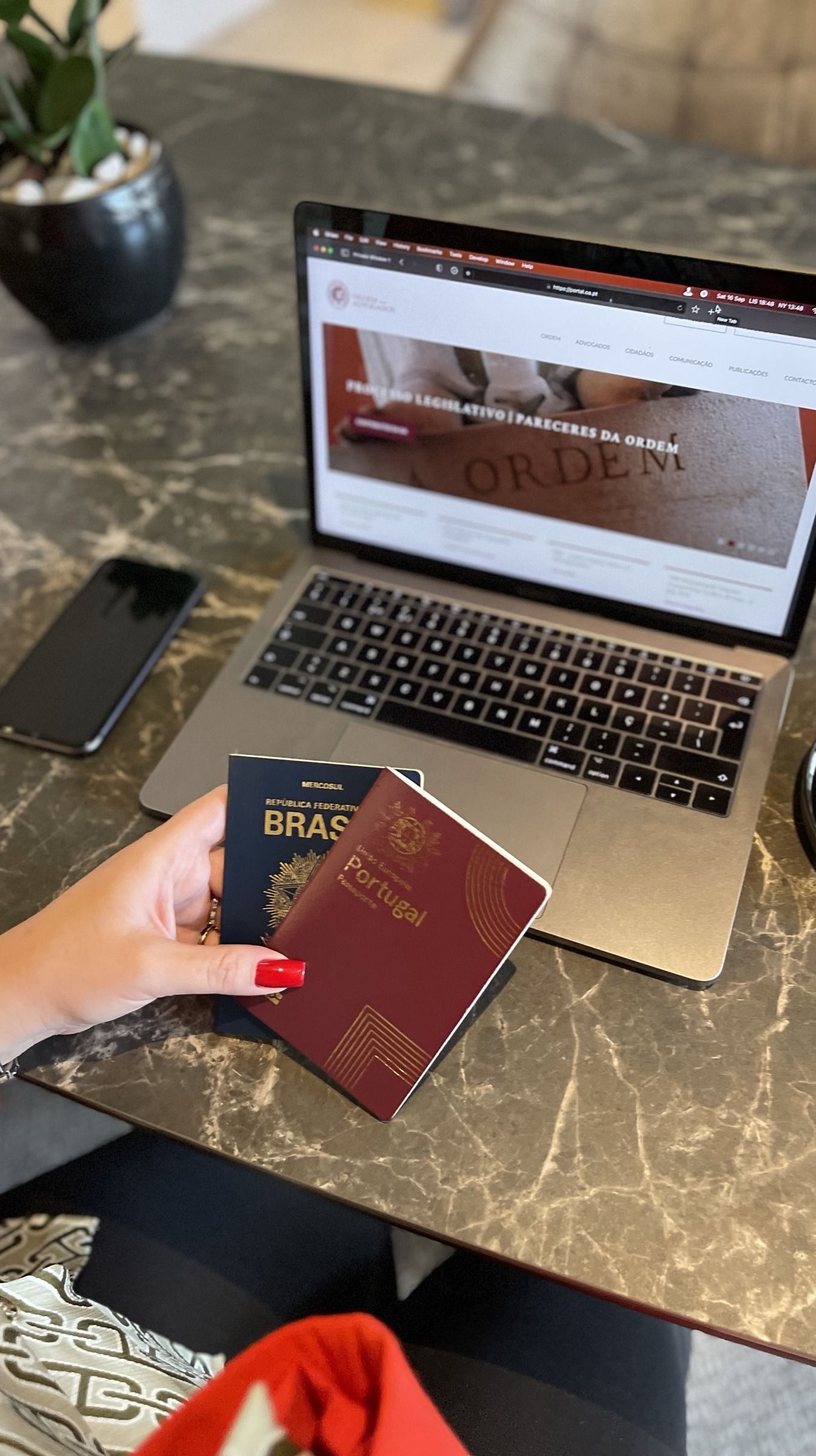 Uma mão segura um passaporte brasileiro e um passaporte português, em frente a um portátil aberto no portal da Ordem dos Advogados